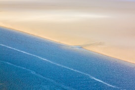A tranquil scene of a coastline with a clear demarcation between the sandy beach and the calm blue ocean. Soft gradients of sand meet with blue rippling water, creating a serene and minimalistic composition.