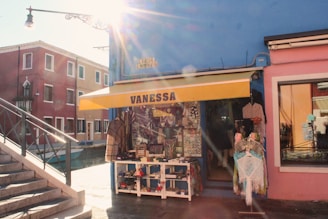 A sunlit street scene features a small shop with a yellow awning displaying the name 'Vanessa'. The shop is painted blue and surrounded by colorful buildings. Various clothing and trinkets are displayed outside, and a canal with a blue boat runs alongside the street.