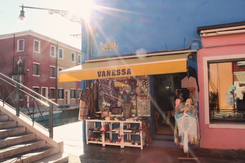 A sunlit street scene features a small shop with a yellow awning displaying the name 'Vanessa'. The shop is painted blue and surrounded by colorful buildings. Various clothing and trinkets are displayed outside, and a canal with a blue boat runs alongside the street.