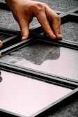 A hand is placing or adjusting a glass panel within a black metal grid structure on a gray, textured surface. The reflective quality of the glass shows faint reflections.