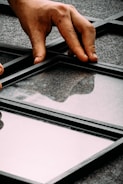 A hand is placing or adjusting a glass panel within a black metal grid structure on a gray, textured surface. The reflective quality of the glass shows faint reflections.