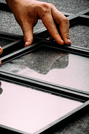 A hand is placing or adjusting a glass panel within a black metal grid structure on a gray, textured surface. The reflective quality of the glass shows faint reflections.