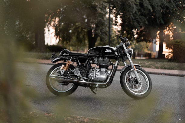 A classic Royal Enfield motorcycle is parked on a road surrounded by lush green trees. The motorcycle has a shiny black body with chrome details and is positioned at an angle, highlighting its sleek design. The background is slightly blurred, emphasizing the motorcycle as the focal point.