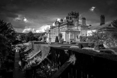 A grand estate with intricate classical architecture stands majestically against a dramatic sky. Beautifully manicured gardens with carefully trimmed hedges and pathways lead up to the structure. The scene is serene with a few people strolling along the garden paths. The atmosphere distinctly captures a sense of elegance and history.