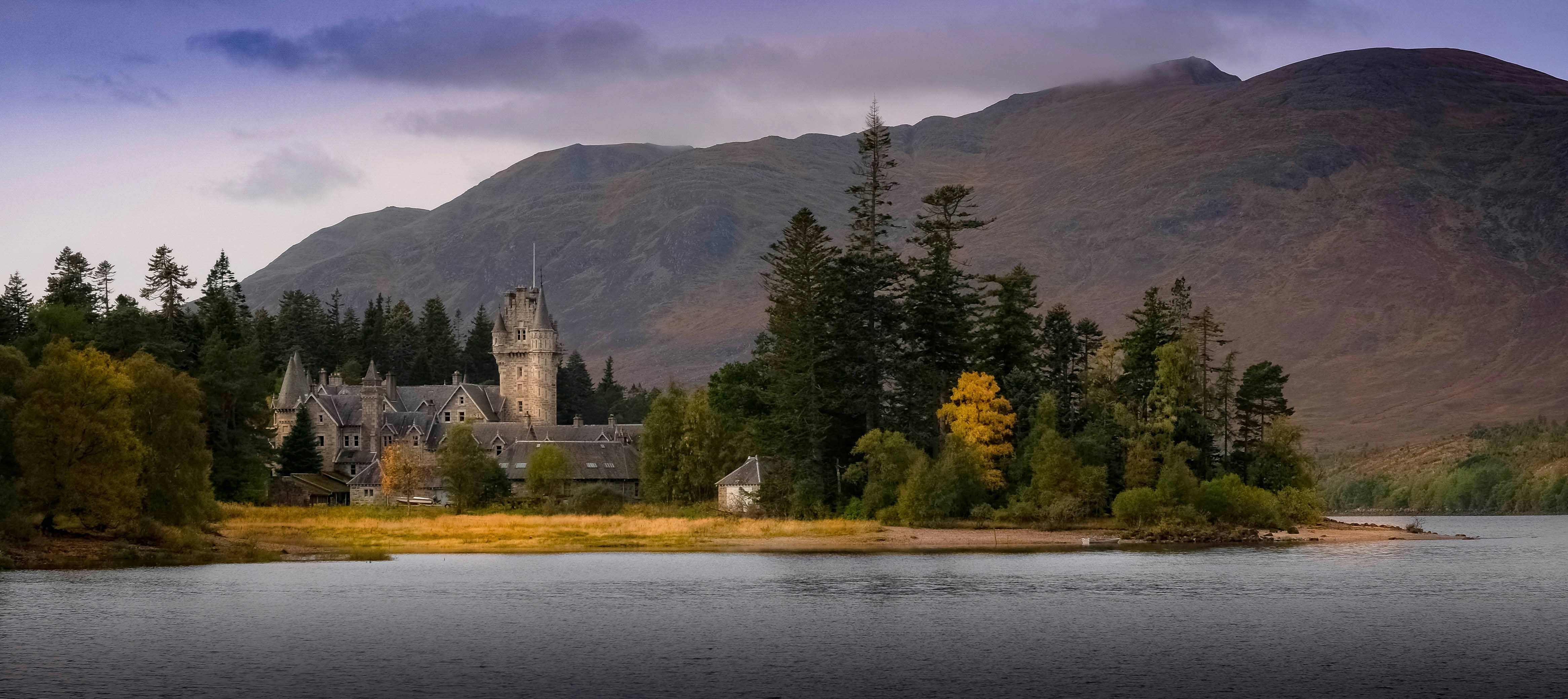a castle on a hill by a lake with trees and mountains in the background