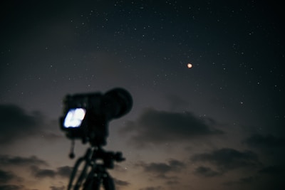 A dimly lit night sky with a visible lunar eclipse. A camera on a tripod is prominently silhouetted against the backdrop of scattered stars and faint clouds.