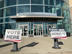 "Vote Here" signs standing outside the glass entrance of a shopping mall