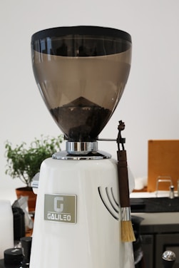 A large coffee grinder with a transparent bean hopper partially filled with coffee beans, sitting on a kitchen counter. The grinder has a sleek white body with chrome accents and a small brush hanging on the side. In the background, there is a potted plant, some appliances, and kitchen items.