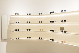 A variety of sunglasses are displayed on curved, backlit shelves in a modern showroom. The shelves are mounted on a light-colored wall, creating a clean and minimalist aesthetic. Different styles and colors of sunglasses are neatly arranged, with some featuring labels or small display signs.