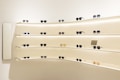 A variety of sunglasses are displayed on curved, backlit shelves in a modern showroom. The shelves are mounted on a light-colored wall, creating a clean and minimalist aesthetic. Different styles and colors of sunglasses are neatly arranged, with some featuring labels or small display signs.