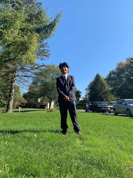 A person stands confidently on a lush green lawn, wearing a formal black suit. The background features a sunny blue sky, with large green trees and a house visible in the distance. Several cars are parked along the driveway.