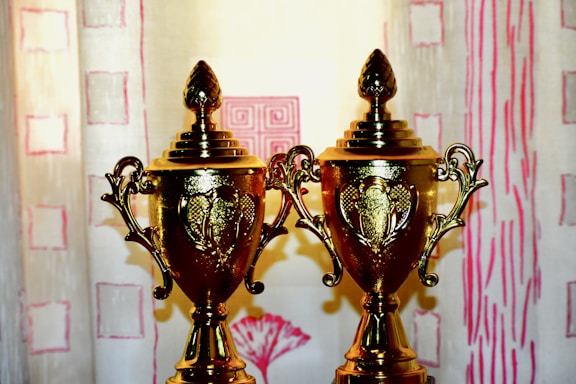Two golden trophies with intricate designs are placed side by side in front of a light-colored patterned background. The trophies have ornate handles and lids with a decorative finial on top.