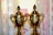 Two golden trophies with intricate designs are placed side by side in front of a light-colored patterned background. The trophies have ornate handles and lids with a decorative finial on top.