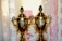 Two golden trophies with intricate designs are placed side by side in front of a light-colored patterned background. The trophies have ornate handles and lids with a decorative finial on top.