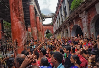 A large crowd of people is gathered in a historic courtyard with tall, weathered red pillars and worn buildings. The individuals are wearing colorful clothing and appear to be enthusiastically participating in an event, with many raising their hands in the air. The atmosphere is lively and vibrant.