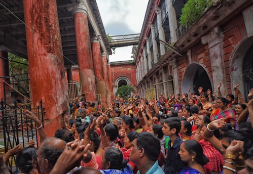 A large crowd of people is gathered in a historic courtyard with tall, weathered red pillars and worn buildings. The individuals are wearing colorful clothing and appear to be enthusiastically participating in an event, with many raising their hands in the air. The atmosphere is lively and vibrant.