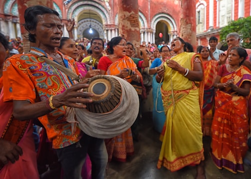 A lively, colorful group of people engaged in a cultural or religious event. The central figure is playing a traditional drum while others around him sing and dance passionately. The background features ornate, historical architecture.