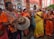 A lively, colorful group of people engaged in a cultural or religious event. The central figure is playing a traditional drum while others around him sing and dance passionately. The background features ornate, historical architecture.