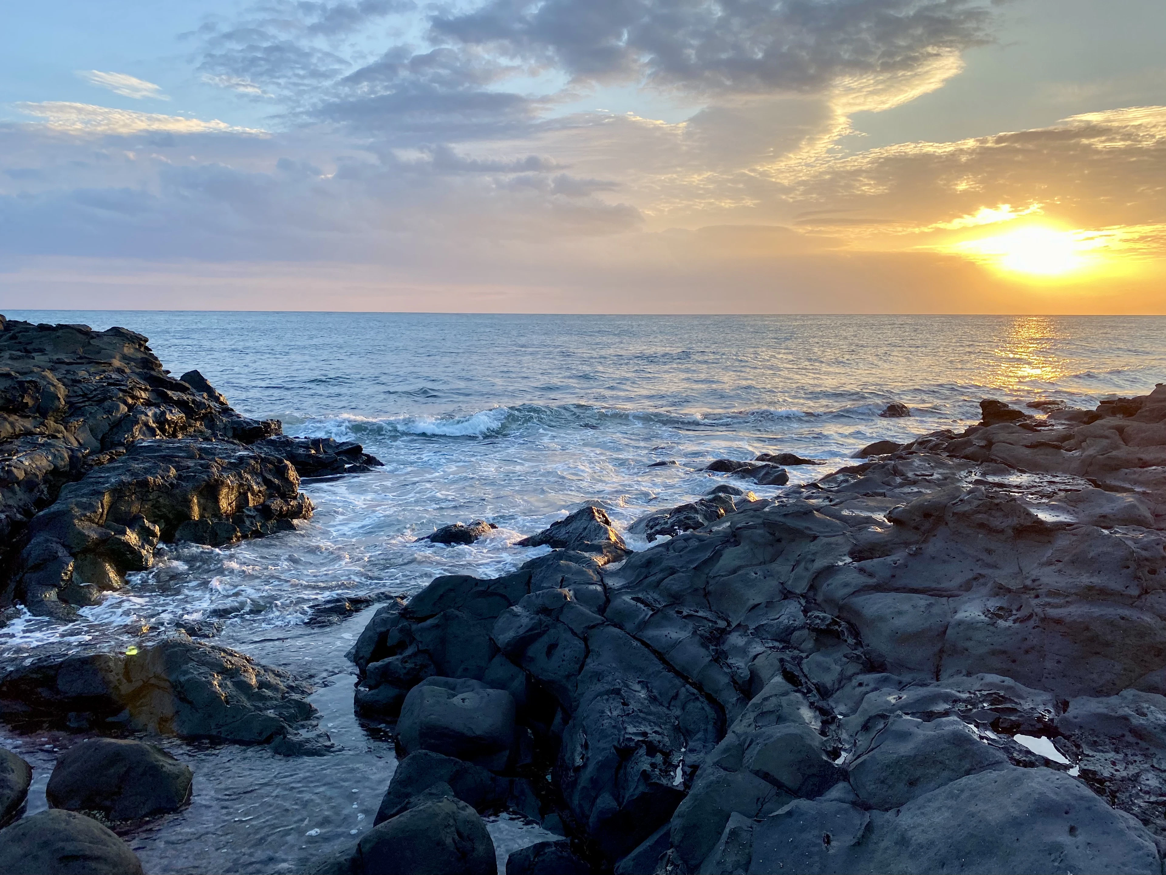 Golden sunlight glows low over the ocean beyond dark lava rocks and breaking waves.