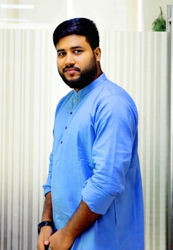 A man with a beard is wearing a light blue traditional kurta and has his hands together in front. The background features a vertical striped pattern.