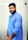 A man with a beard is wearing a light blue traditional kurta and has his hands together in front. The background features a vertical striped pattern.