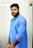 A man with a beard is wearing a light blue traditional kurta and has his hands together in front. The background features a vertical striped pattern.