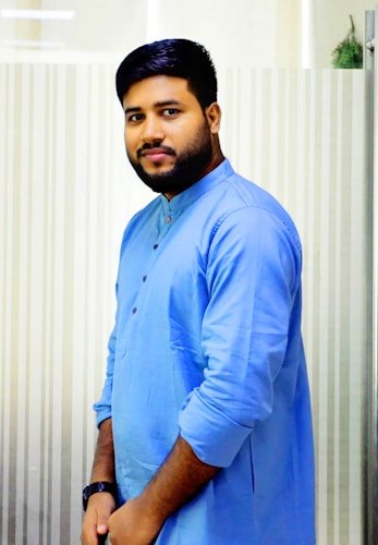 A man with a beard is wearing a light blue traditional kurta and has his hands together in front. The background features a vertical striped pattern.