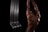 A muscular person with a tattooed back is stretching their arms above their head. The lighting creates highlights on the skin and the black background emphasizes the subject. On the left side, there are elongated metallic structures resembling pipes.
