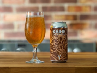 A glass filled with a golden, bubbly beverage is placed on a wooden surface next to a can with an artistic design labeled 'KAIHE DDH CHINOOK IPA'. The background features a blurred brick wall.