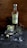 A bottle of Prairie organic gin is placed on a rustic wooden surface next to a glass filled with gin, ice, and lime wedges. Around the glass, there are scattered ice cubes and a small plant with spiky leaves, along with a small animal skull.