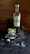 A bottle of Prairie organic gin is placed on a rustic wooden surface next to a glass filled with gin, ice, and lime wedges. Around the glass, there are scattered ice cubes and a small plant with spiky leaves, along with a small animal skull.