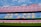 A large empty football stadium with rows of red and blue seats. The central section of seats forms the words 'MES QUE UN CLUB' in yellow letters. There are advertising banners for various brands at the top and bottom of the stands. The stadium field is visible in the foreground under a clear, partly cloudy sky.