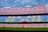 A large empty football stadium with rows of red and blue seats. The central section of seats forms the words 'MES QUE UN CLUB' in yellow letters. There are advertising banners for various brands at the top and bottom of the stands. The stadium field is visible in the foreground under a clear, partly cloudy sky.