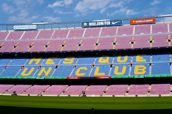 A large empty football stadium with rows of red and blue seats. The central section of seats forms the words 'MES QUE UN CLUB' in yellow letters. There are advertising banners for various brands at the top and bottom of the stands. The stadium field is visible in the foreground under a clear, partly cloudy sky.