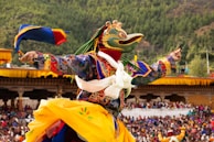 A vibrant festival scene with a person wearing a colorful, elaborately decorated costume and a large bird-like mask, performing a traditional dance in an outdoor setting. The background is filled with spectators and lush green trees covering the hillside.