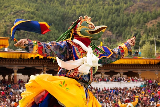 A vibrant festival scene with a person wearing a colorful, elaborately decorated costume and a large bird-like mask, performing a traditional dance in an outdoor setting. The background is filled with spectators and lush green trees covering the hillside.