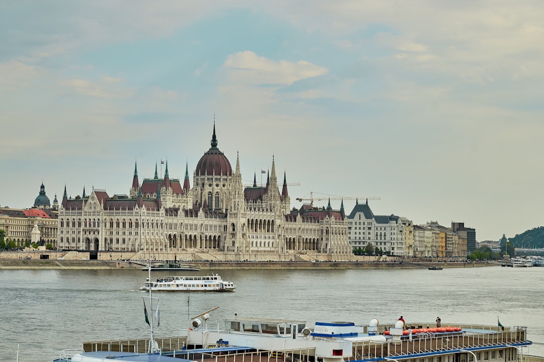 a large building with towers and a domed roof by a body of water with Hungarian Parliament Building in the background,