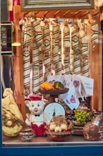 A display window featuring an assortment of sausages hanging in the background, adorned with colorful packaging. In the foreground, there is a playful ceramic pig dressed in a red and white outfit. The display includes various ceramic bowls filled with items like garlic, grapes, peppers, and a large cheese. A vintage scale, decorated with fruit and vegetables, is prominently displayed.