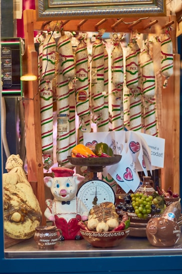 A display window featuring an assortment of sausages hanging in the background, adorned with colorful packaging. In the foreground, there is a playful ceramic pig dressed in a red and white outfit. The display includes various ceramic bowls filled with items like garlic, grapes, peppers, and a large cheese. A vintage scale, decorated with fruit and vegetables, is prominently displayed.
