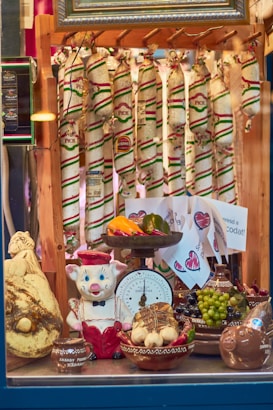 A display window featuring an assortment of sausages hanging in the background, adorned with colorful packaging. In the foreground, there is a playful ceramic pig dressed in a red and white outfit. The display includes various ceramic bowls filled with items like garlic, grapes, peppers, and a large cheese. A vintage scale, decorated with fruit and vegetables, is prominently displayed.