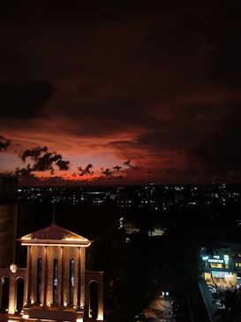 A dramatic sunset sky filled with deep orange and red hues, with scattered clouds creating a striking silhouette against the skyline. Below, an illuminated building with a classical facade stands out, adorned with glowing lights and a visible clock. In the background, city lights are visible, hinting at an urban environment beyond the foreground architecture.