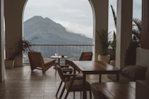 A serene terrace with wooden chairs and tables overlooks a majestic mountain landscape. Potted plants add a touch of greenery, and there is a small table with cups, suggesting a relaxing setting.