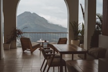 A serene terrace with wooden chairs and tables overlooks a majestic mountain landscape. Potted plants add a touch of greenery, and there is a small table with cups, suggesting a relaxing setting.