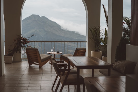 A serene terrace with wooden chairs and tables overlooks a majestic mountain landscape. Potted plants add a touch of greenery, and there is a small table with cups, suggesting a relaxing setting.