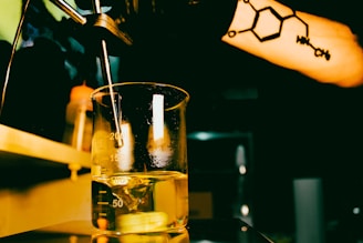 A beaker filled with a yellow liquid sits under a spout in a dimly lit room. A person's arm with a tattoo of a chemical structure is visible in the foreground. The setting appears to be a laboratory with additional equipment and bottles in the background.