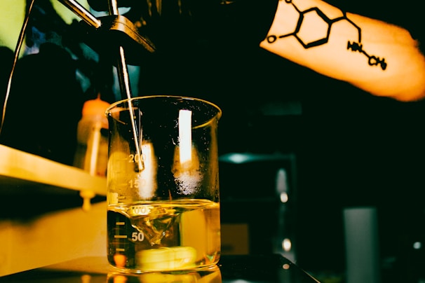 A beaker filled with a yellow liquid sits under a spout in a dimly lit room. A person's arm with a tattoo of a chemical structure is visible in the foreground. The setting appears to be a laboratory with additional equipment and bottles in the background.
