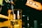 A beaker filled with a yellow liquid sits under a spout in a dimly lit room. A person's arm with a tattoo of a chemical structure is visible in the foreground. The setting appears to be a laboratory with additional equipment and bottles in the background.