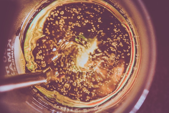 A close-up view of a swirling liquid with numerous small bubbles, possibly carbonation or effervescence, inside a circular glass container. The lighting gives the appearance of golden colors, and a metallic rod is partially submerged in the liquid.