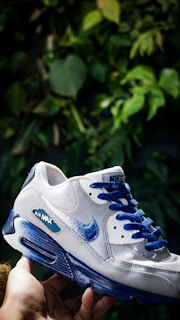 A white sneaker with blue accents is being held in a hand against a background of green leaves. The sneaker features the branding 'Air Max' and the Nike logo in blue.