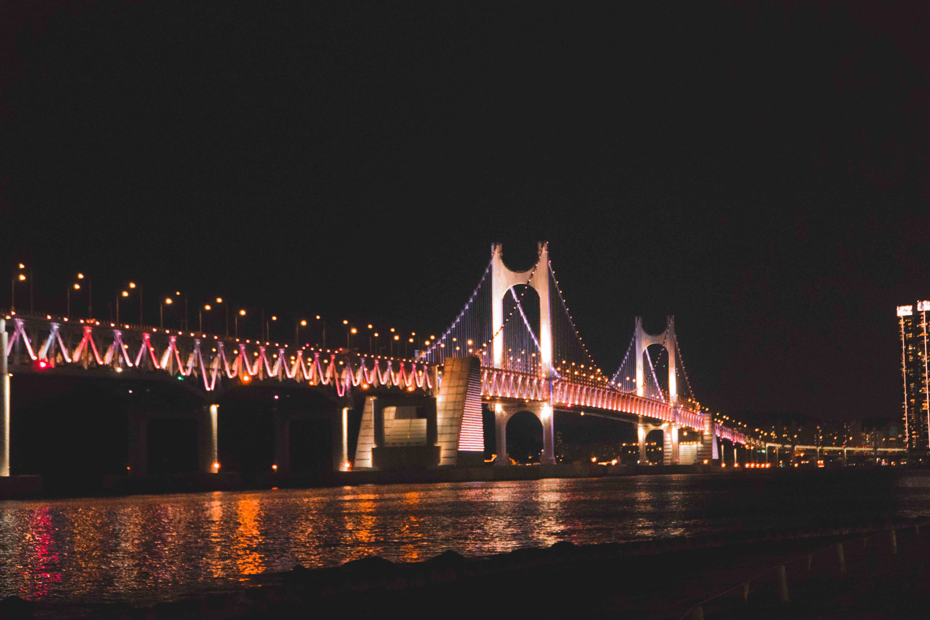 A large illuminated suspension bridge glows red and white over Busan's bay at night, lights reflected in the water.